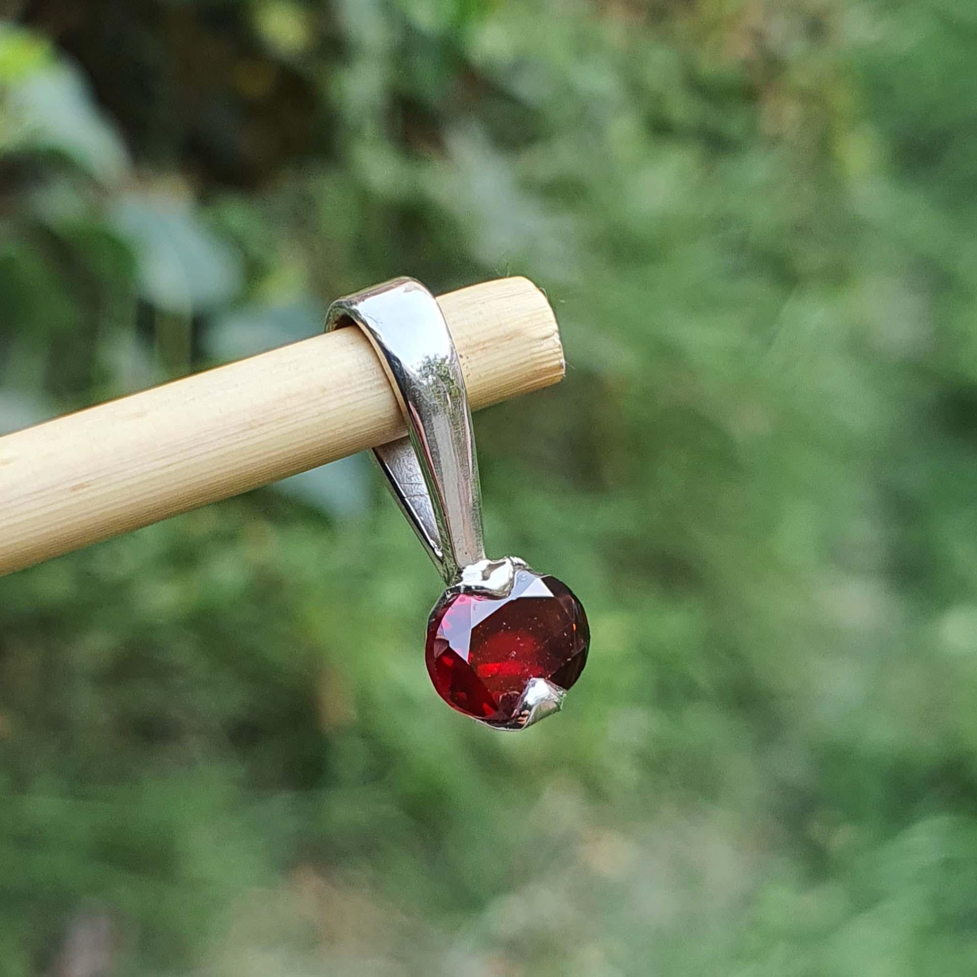 Silver pendant with a red gemstone held by a wooden stick against a blurred green background