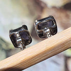 Pair of silver square earrings with purple stones on a wooden stick against a blurred natural background