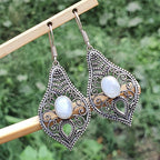 Ornate earrings with moonstone stones on a wooden stick against a green foliage background