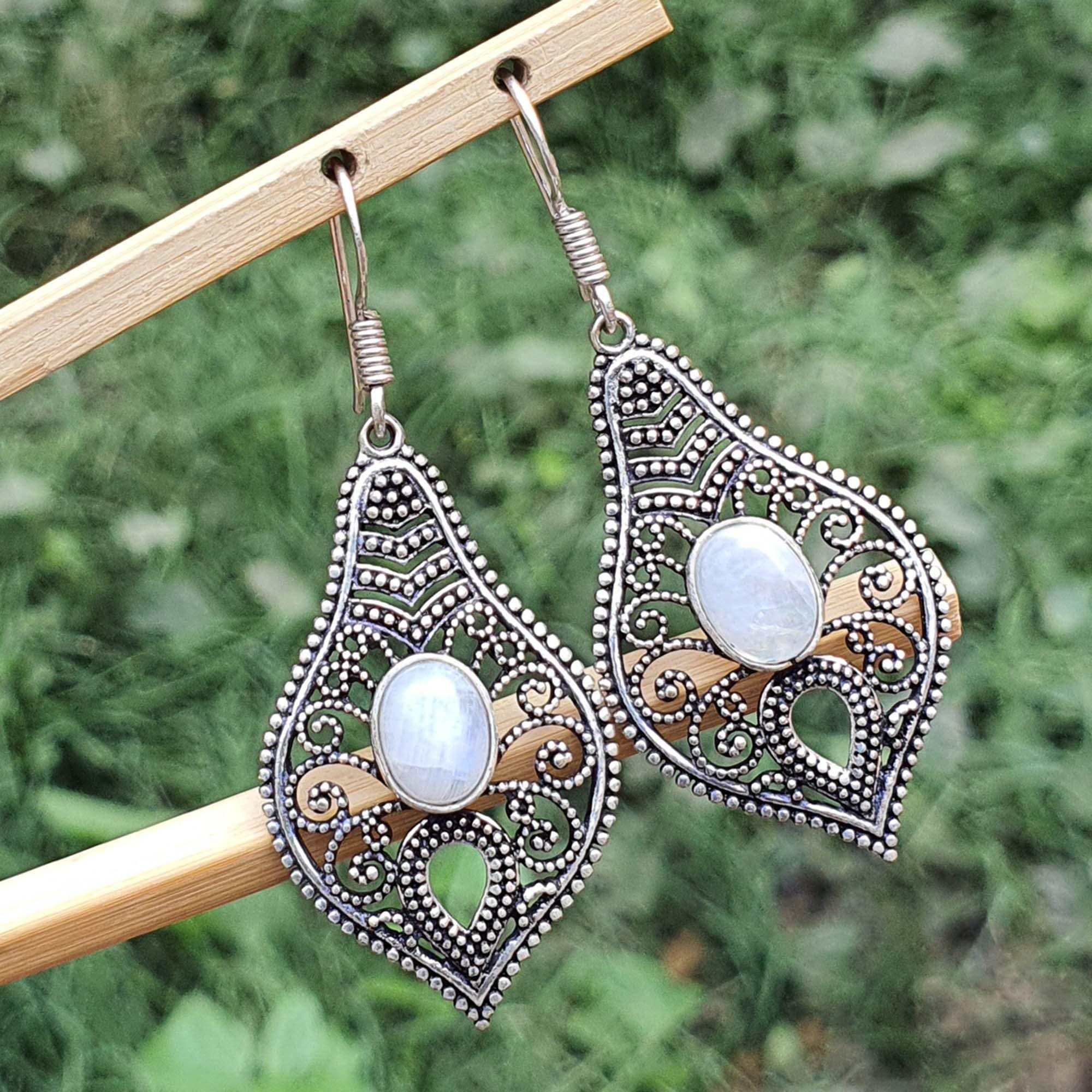 Ornate earrings with moonstone stones on a wooden stick against a green foliage background