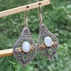 Ornate silver and gold earrings with white stones on a wooden stick against a green foliage background.