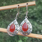 Teardrop-shaped earrings with red stones and silver filigree on a wooden stick against a blurred green background.