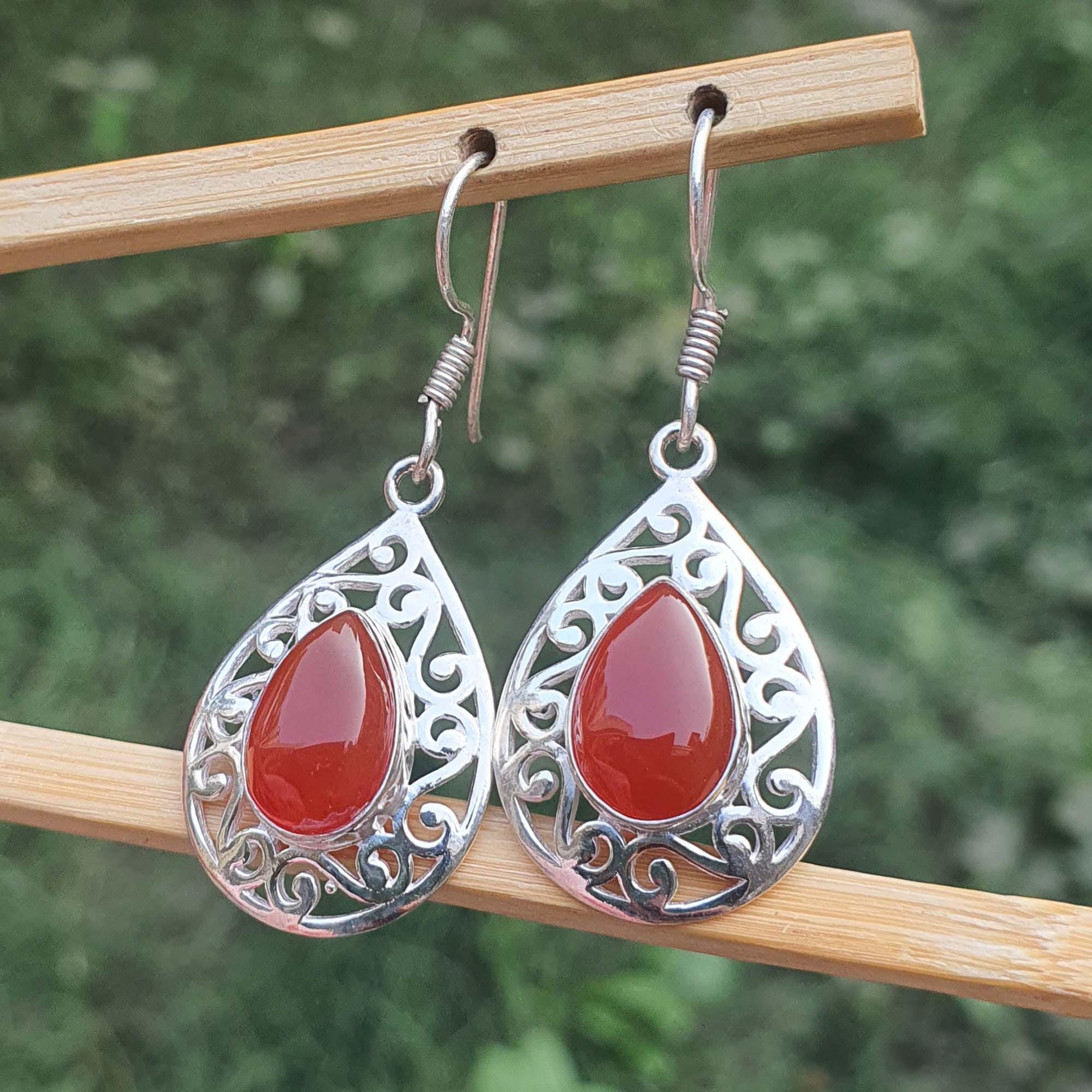 Teardrop-shaped earrings with red stones and silver filigree on a wooden stick against a blurred green background.