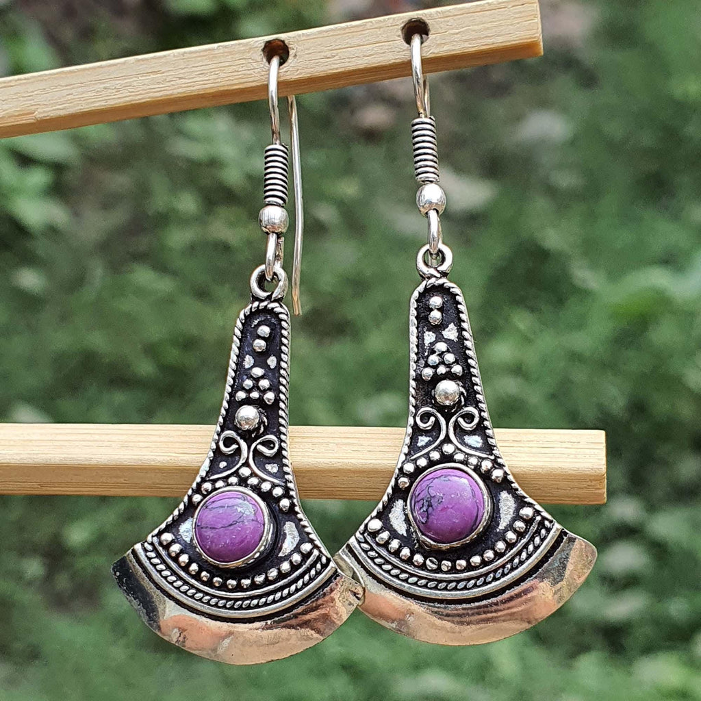 Pair of silver and purple earrings hanging on a wooden stick with a green blurred background.